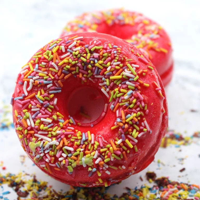 Fruit scented bath bombs shaped like donuts, featuring strawberry and banana colors floating in a bubbly bath scene.