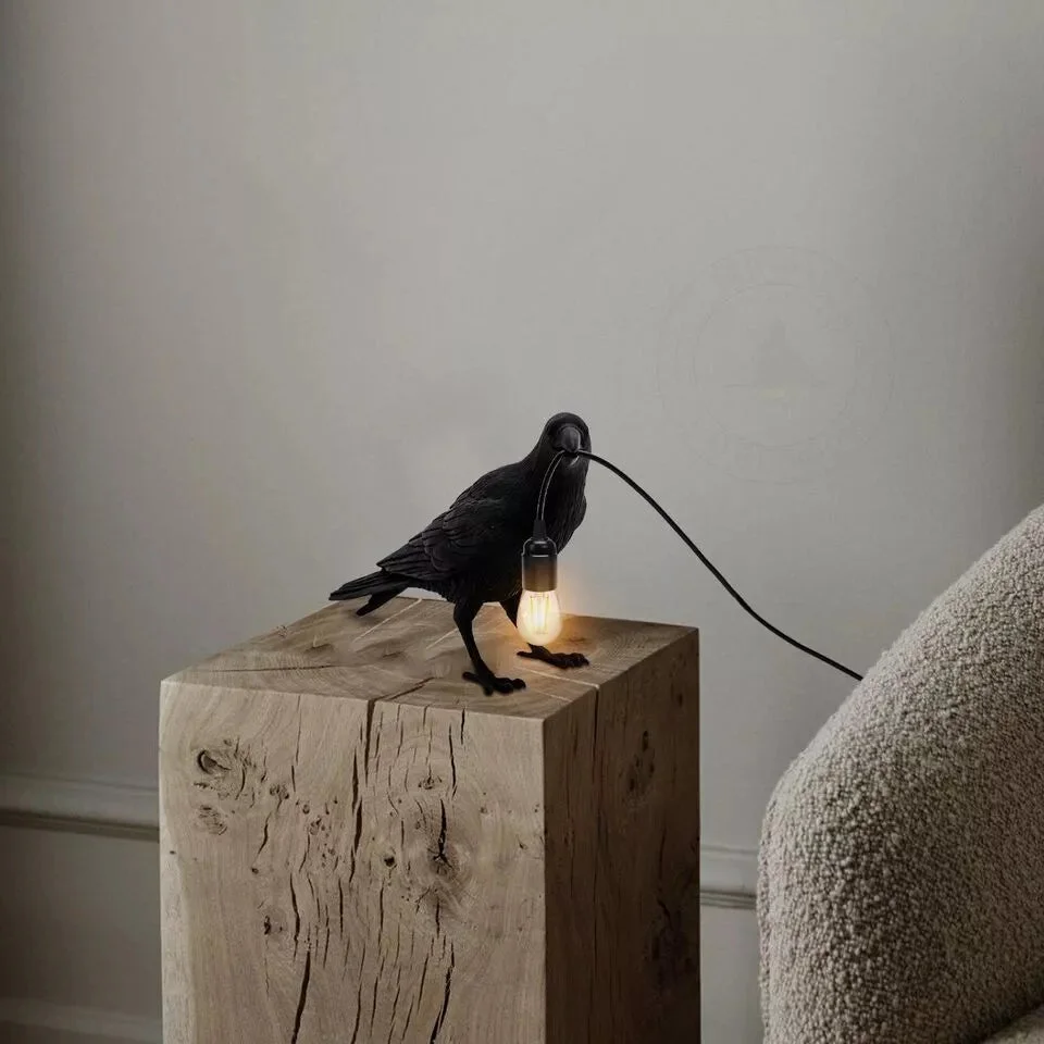 Black resin crow-shaped E27 bulb table lamp shown on a wooden desk with warm light glowing