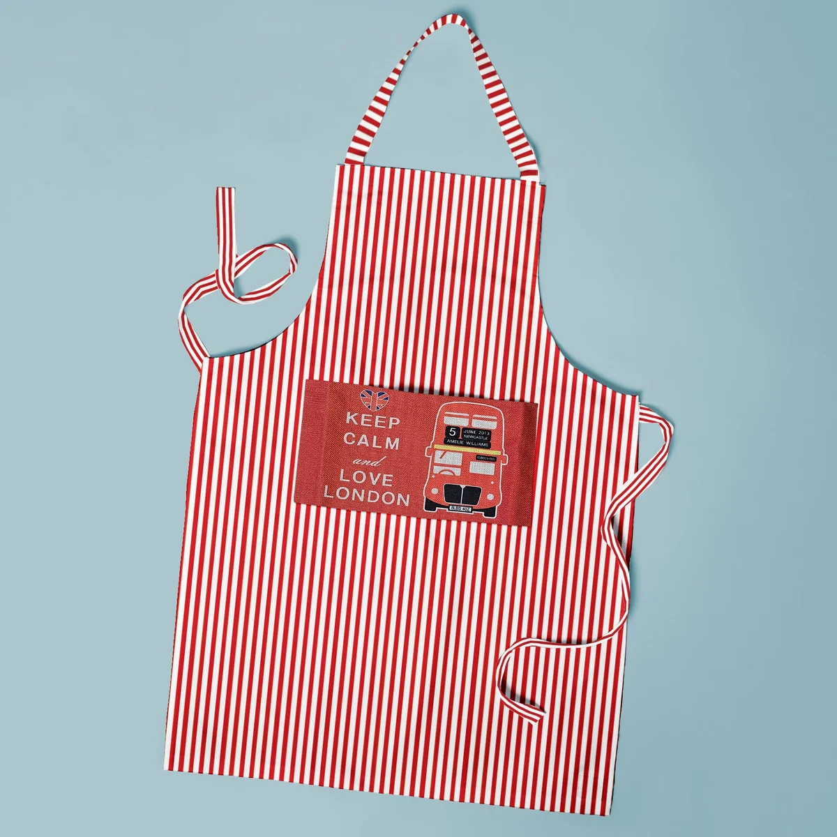 Adjustable neck apron in cotton with a large front pocket and long waist ties, shown flat against a white background