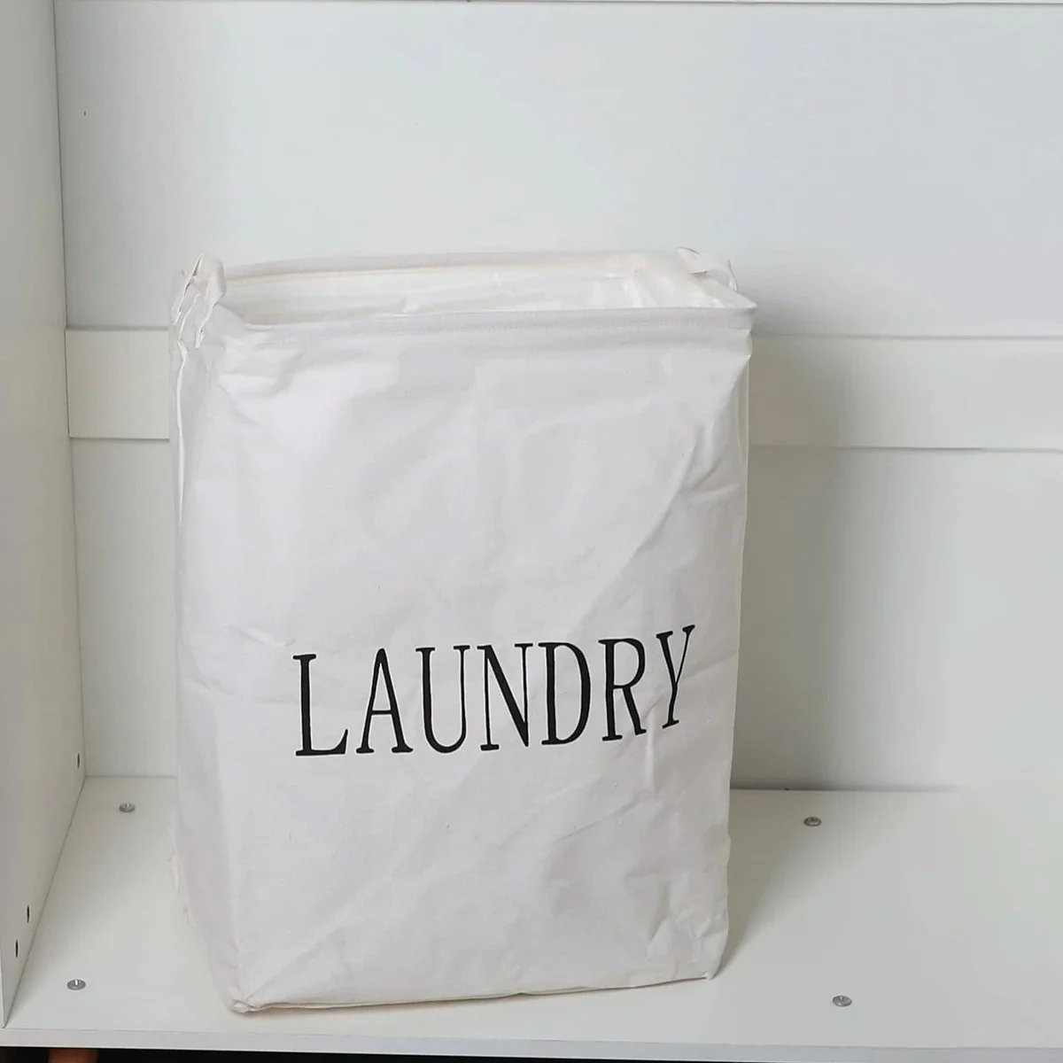 Foldable laundry bag made of linen and cotton, square shape with handles, standing upright in a bedroom setting