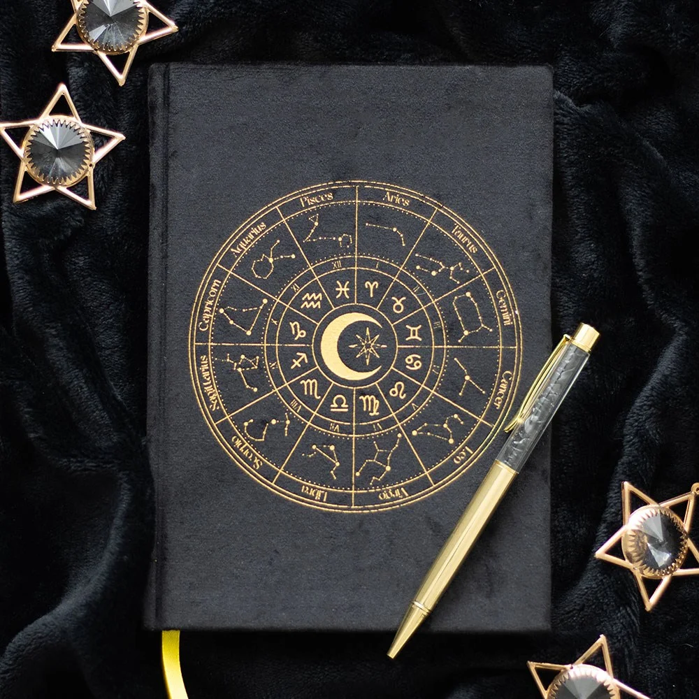 Black velvet astrology journal with gold-edged lined pages and a black obsidian crystal chip ballpoint pen on white background