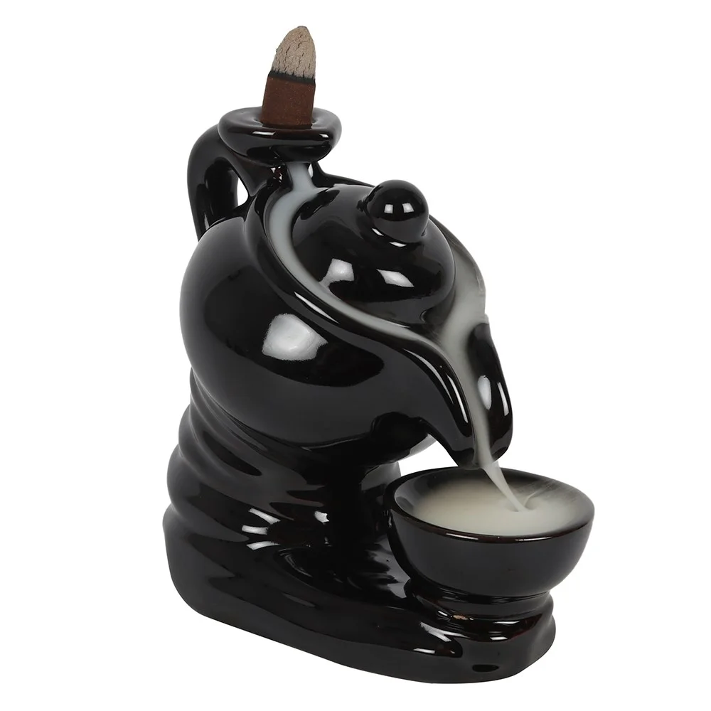 Black ceramic teapot backflow incense burner with smoke flowing from spout into matching teacup below on white background