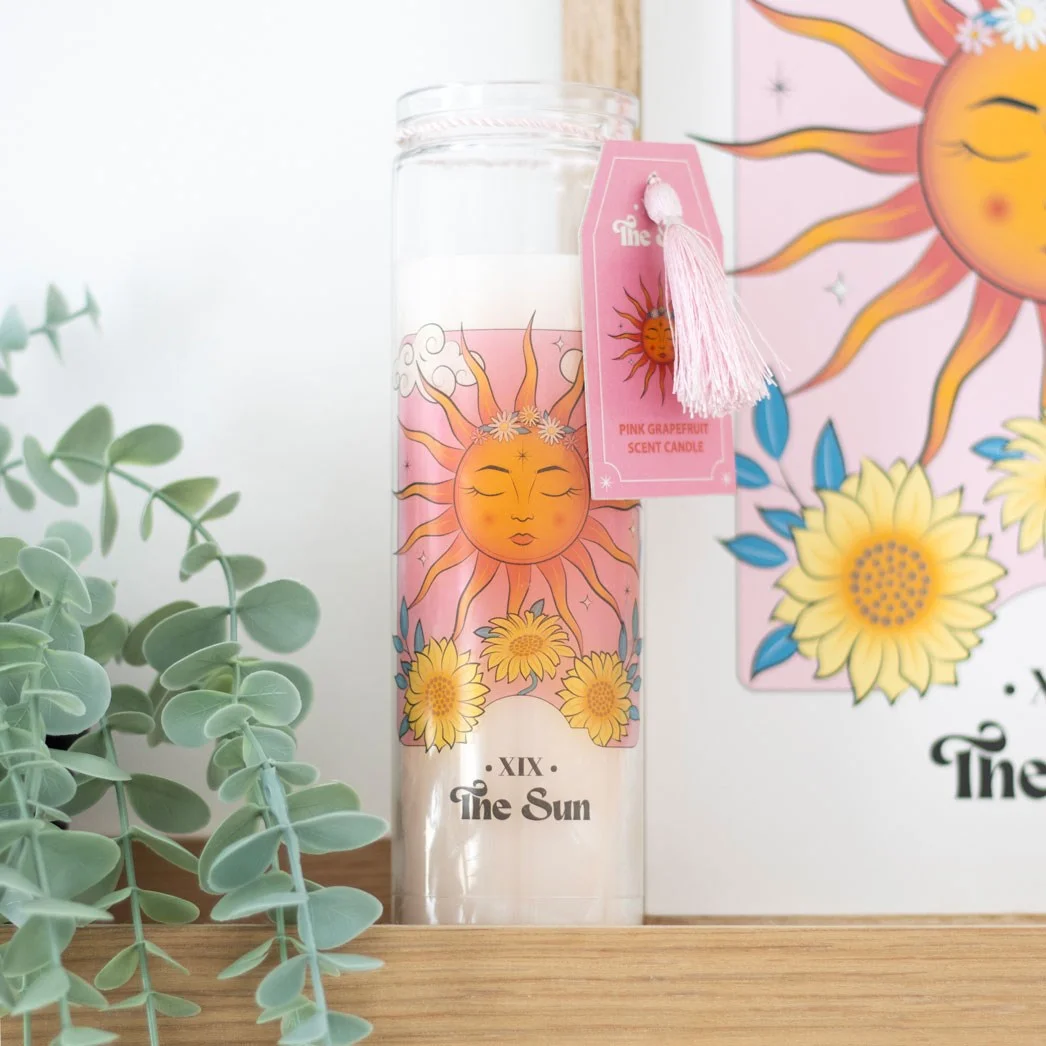 Pink grapefruit candle in a tube with The Sun tarot card design, glowing warmly on a white background