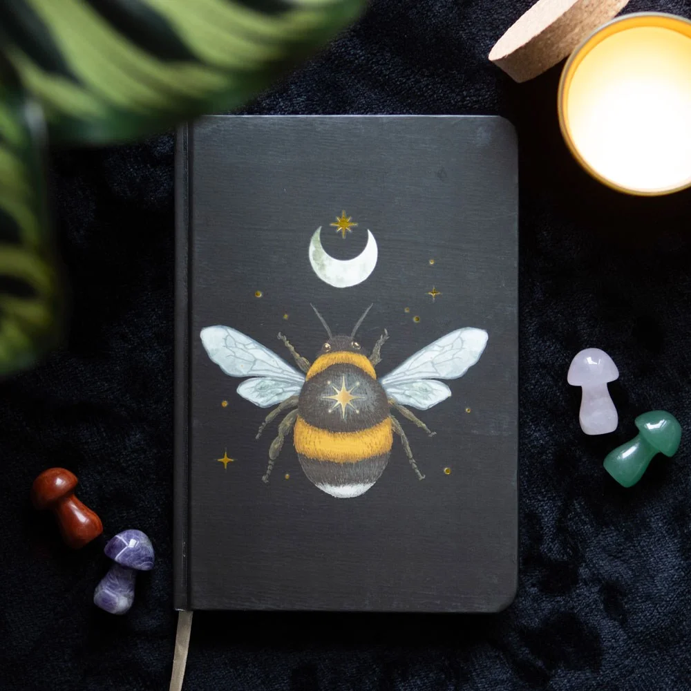 A5 notebook with mystical bee design, gold foil-edged pages, and a ribbon marker on a white background