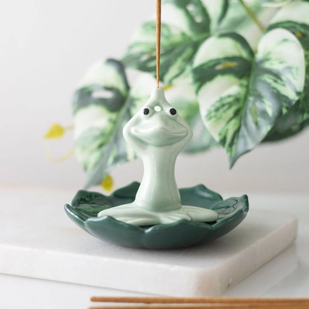 Ceramic incense holder shaped like a yogi frog in sukhasana pose, resting in a green lotus, catching falling ash.