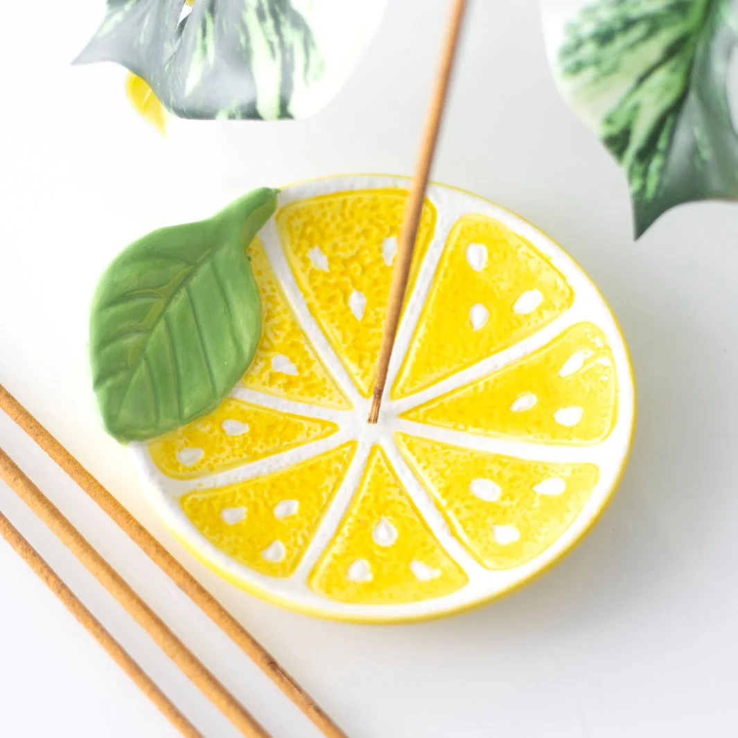 Ceramic lemon slice incense holder in bright yellow, shaped like a lemon wedge, holding a lit incense stick with ash catcher
