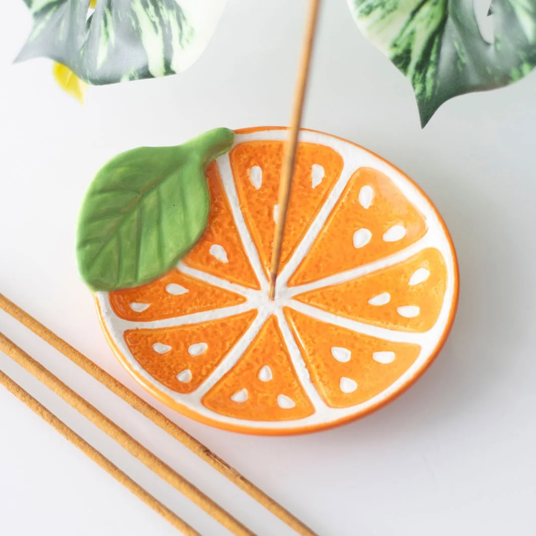 Bright orange slice ceramic incense holder with a lit incense stick, catching ash on a white surface