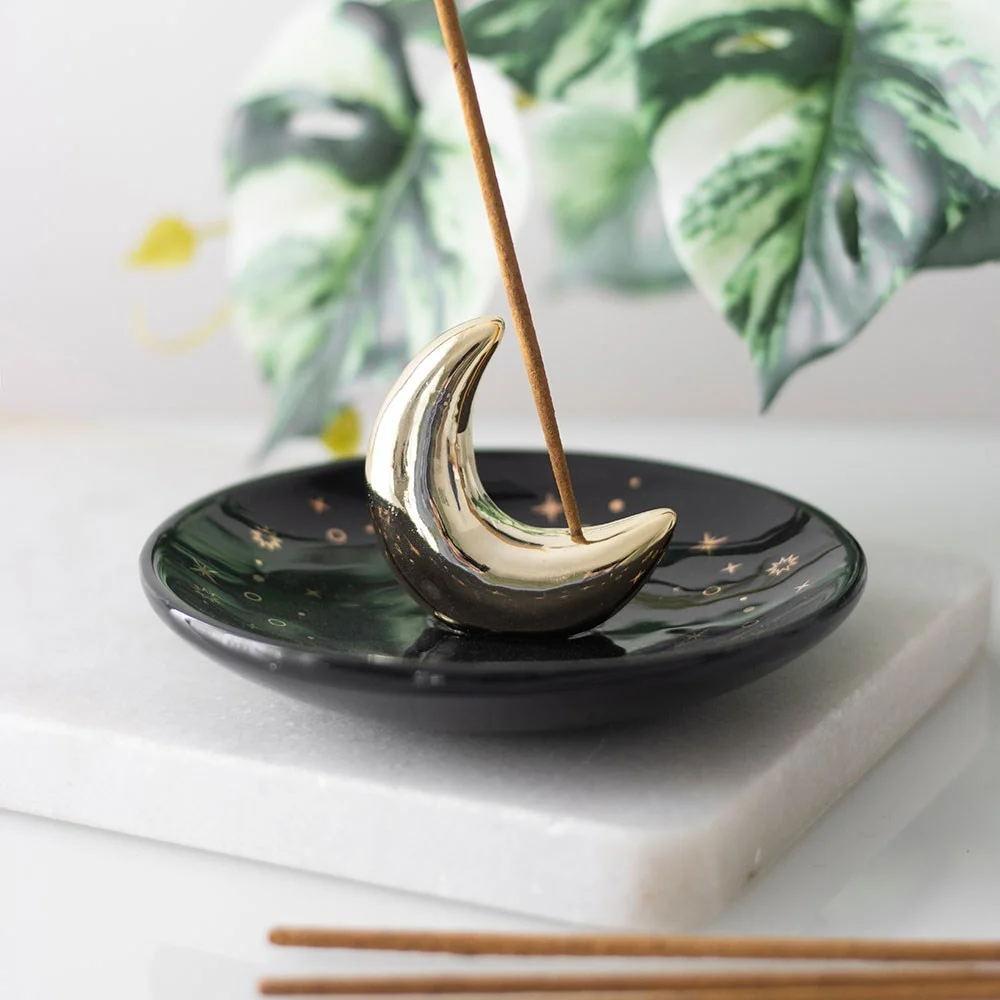 Gold ceramic crescent moon incense holder with a lit incense stick, catching ash on a white background