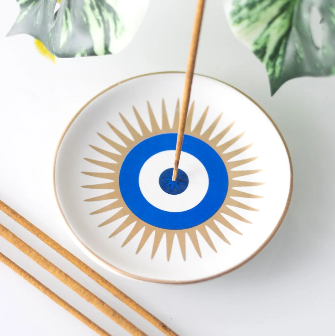 Ceramic incense holder shaped like an all-seeing eye in white, holding a lit incense stick with ash catcher tray