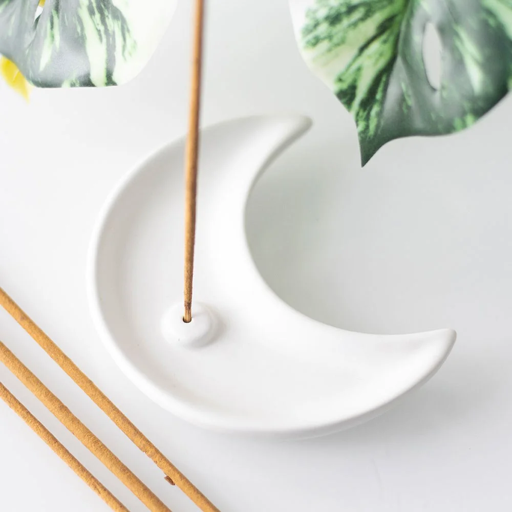 White ceramic incense holder shaped like a crescent moon with incense stick and ash catcher on a white background
