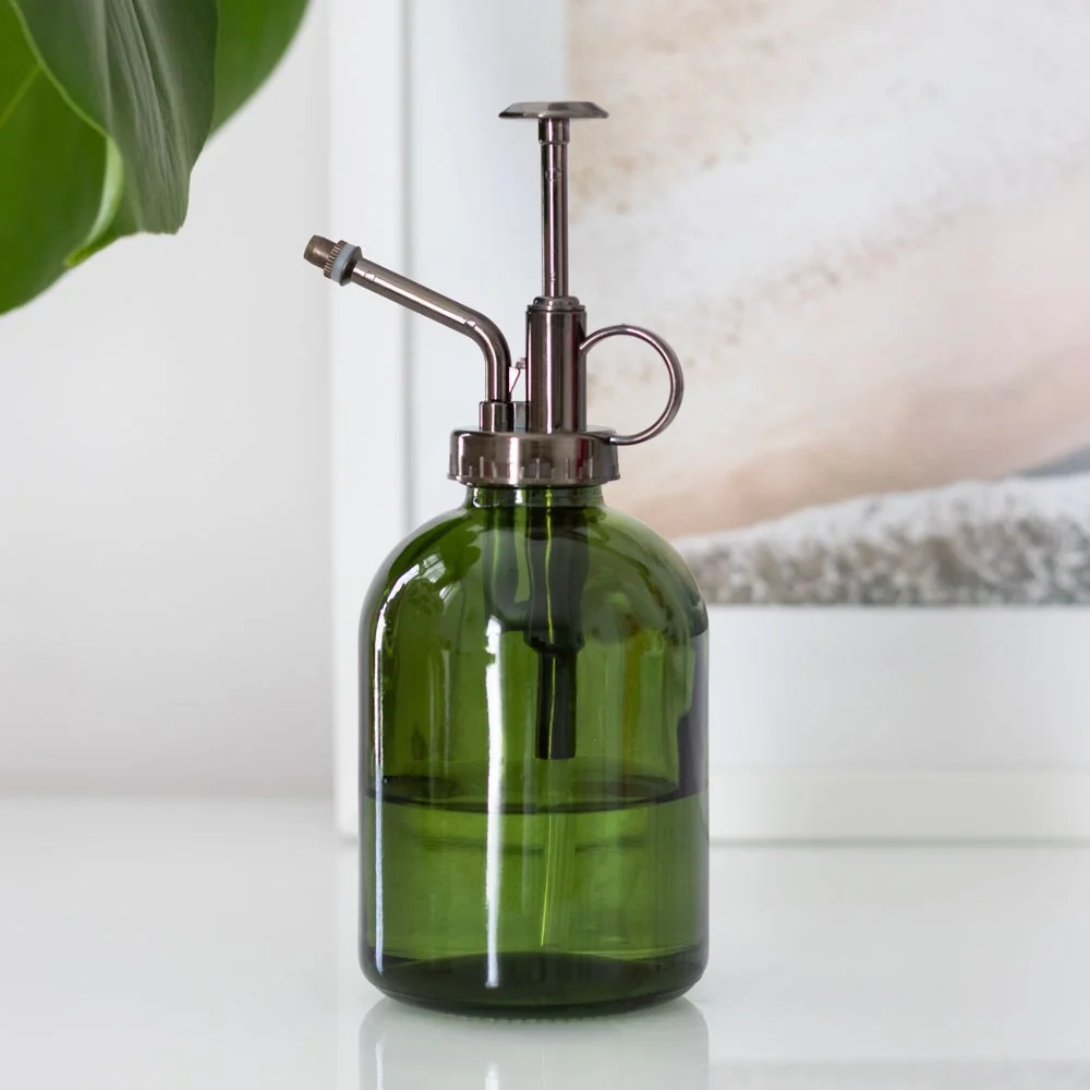 Moss green glass plant mister, minimalist bottle design, glass body with clear stem, faint mist sprayer handle.