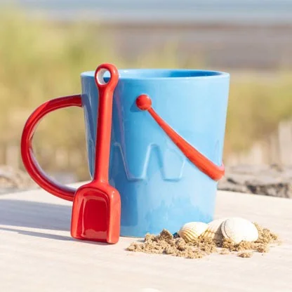Blue ceramic seaside mug shaped like a bucket with a red shovel-shaped spoon inside on a white background