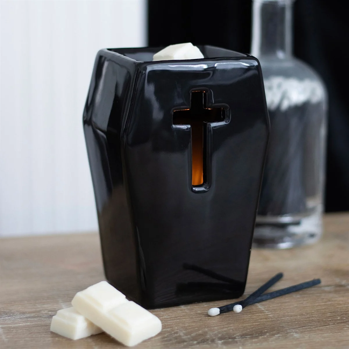 Black coffin oil burner with cut-out cross design glowing from tealight inside, casting ambient light and shadows