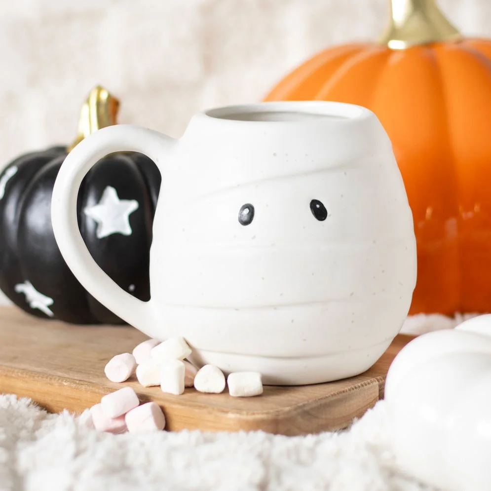 White ceramic mummy shaped mug with wrapped bandage design and black eyes on a plain background