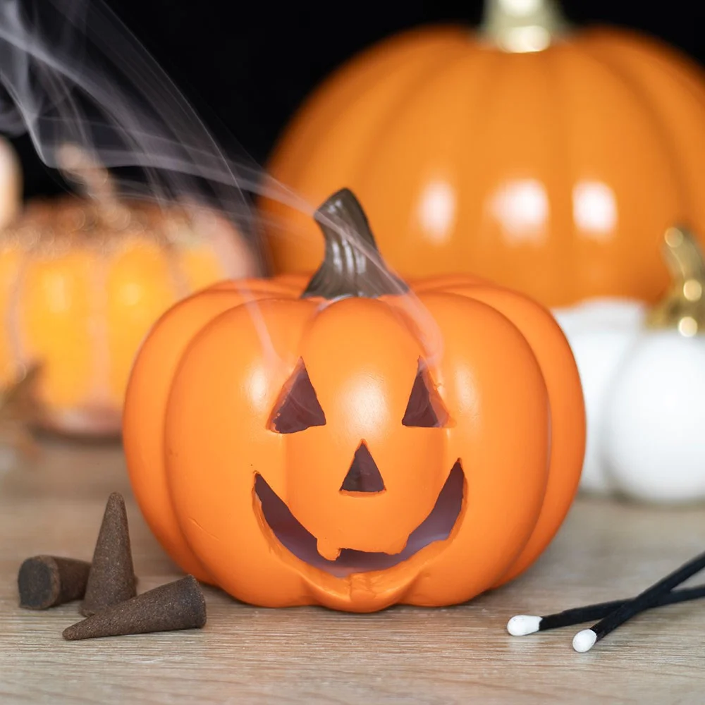 Orange pumpkin incense holder shaped like a jack-o'-lantern with a toothy grin and a lit incense cone inside smoke emerging