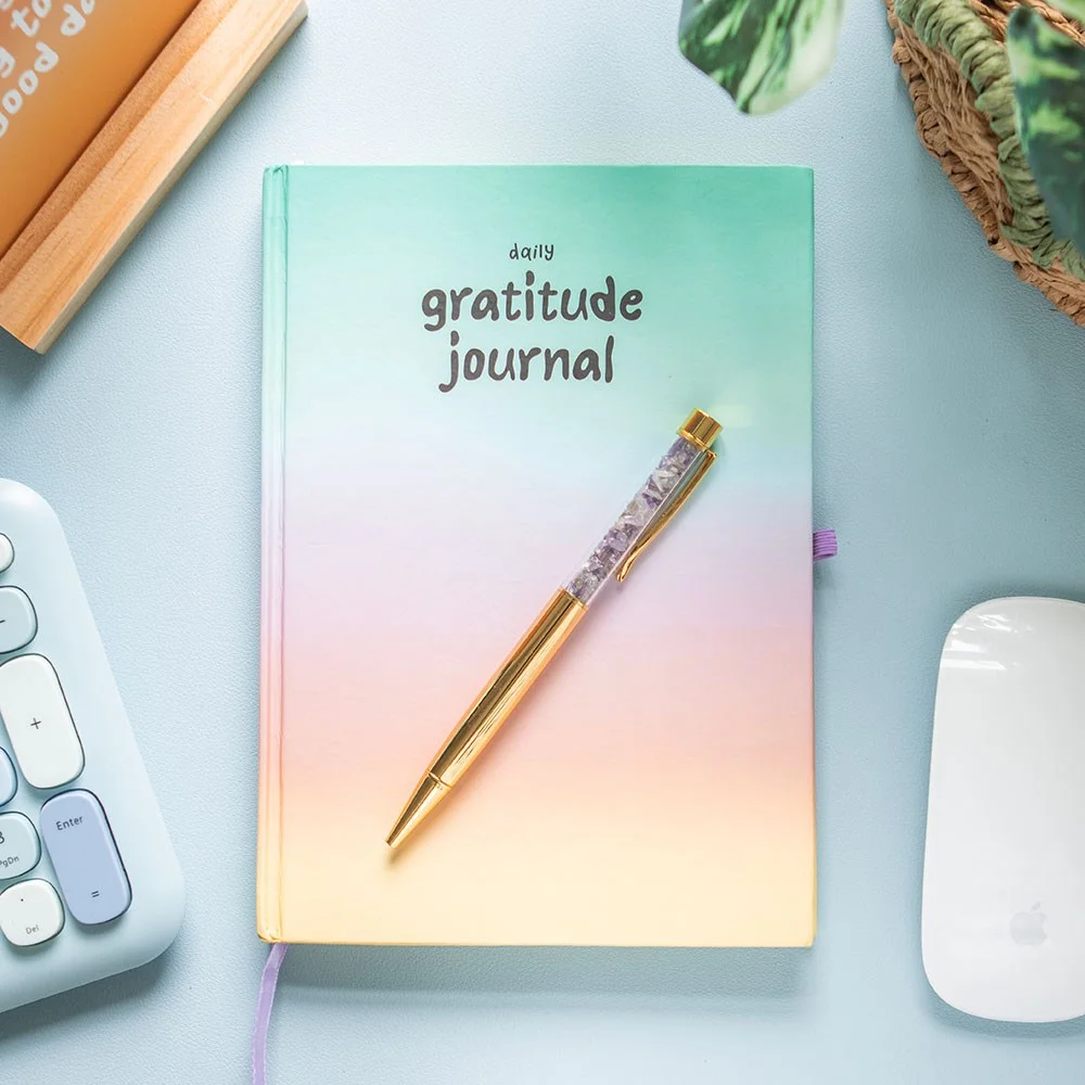 Multicoloured gratitude journal open to lined pages with a matching purple amethyst pen on white background