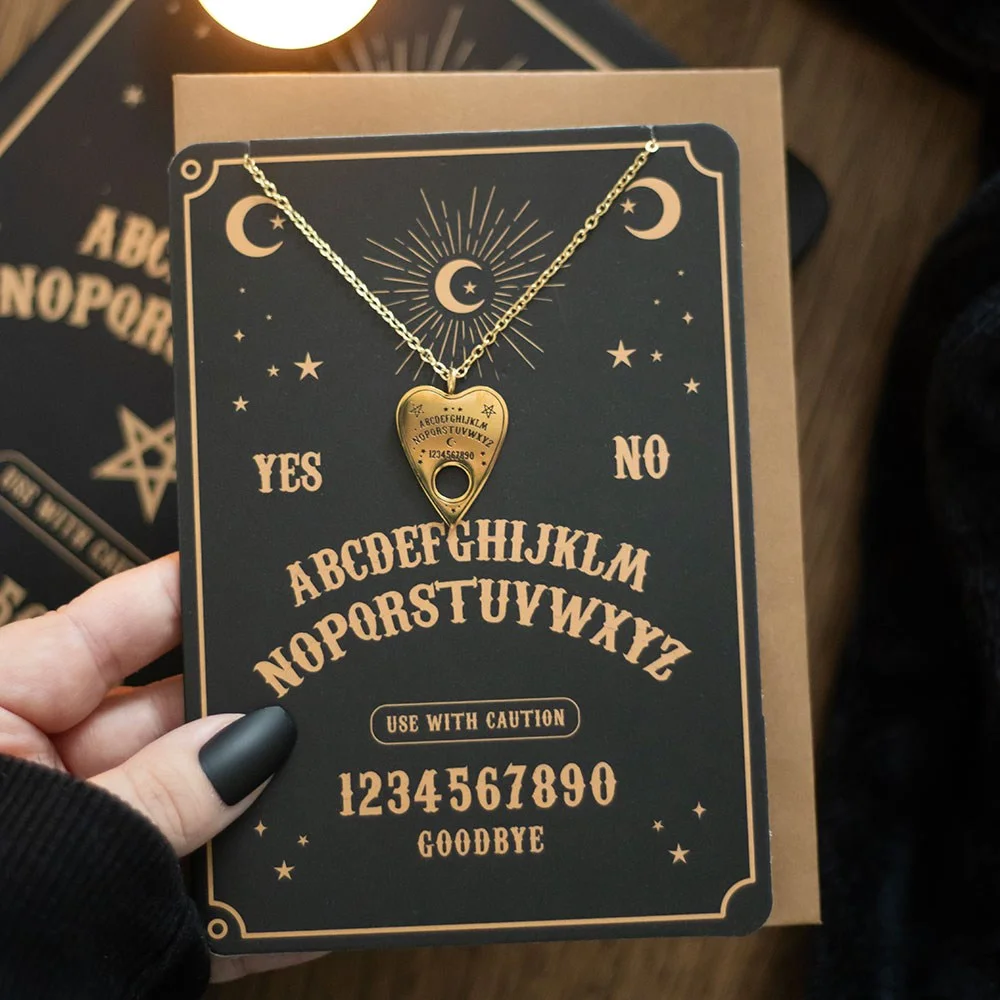Gold-tone talking board necklace with planchette pendant displayed on matching gold-finish greeting card and envelope