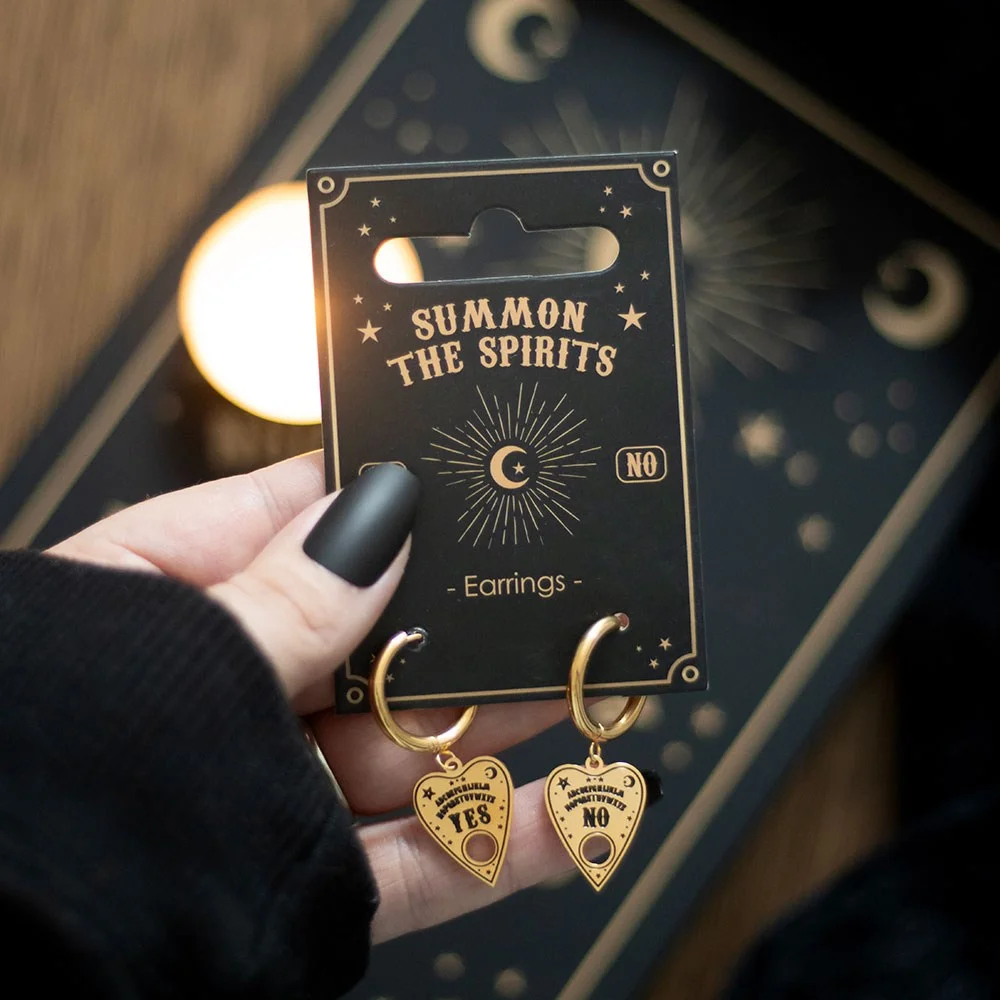 Gold-tone talking board earrings with detailed planchette charms hanging from small hoops on a display card