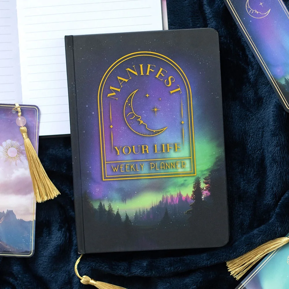 Weekly planner notebook with forest cover, gold foil moon, 'Manifest your life' text, holographic edges, and gold tassel marker