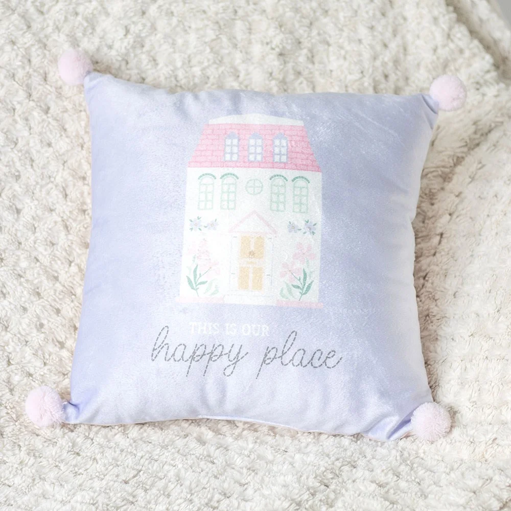 Decorative square cushion with pastel blue and pink house design, pom poms, and "This is our Happy Place" text