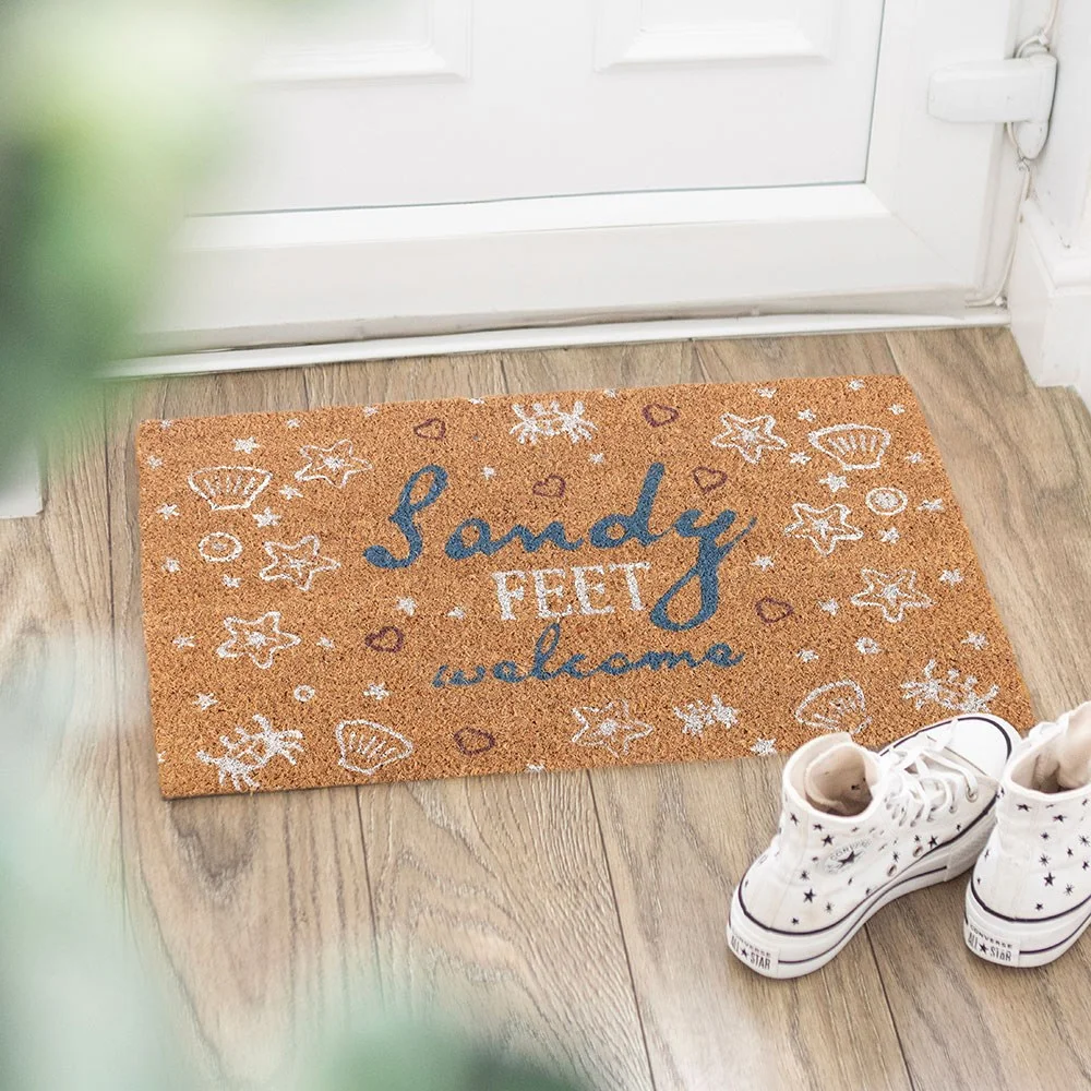 Natural coir doormat with "Sandy feet welcome" text and shell and crab designs in sandy beige tones
