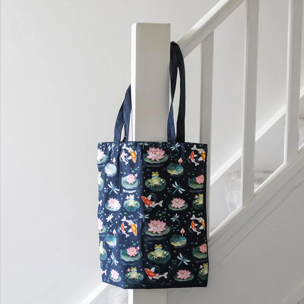 Black polycotton tote bag featuring a pond print with frogs, dragonflies, lily pads, and koi fish design