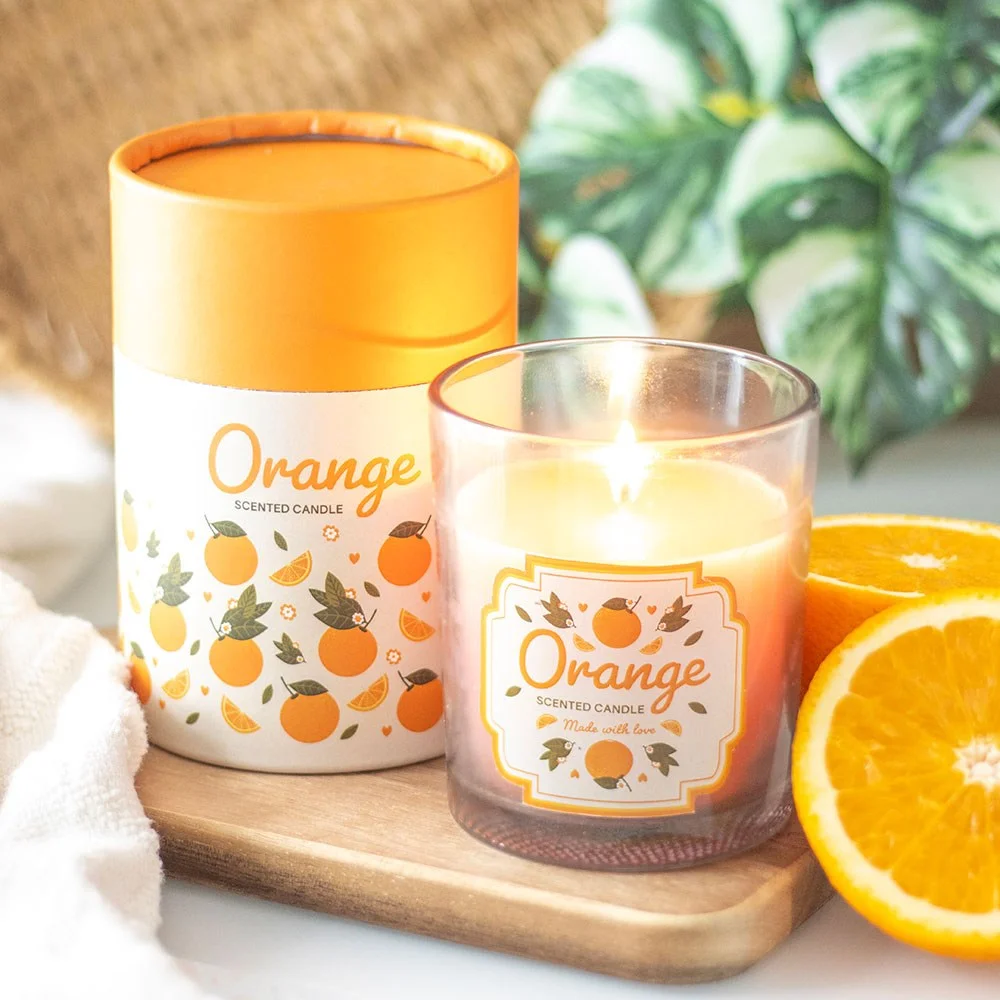 Orange scented candle in a glass holder with orange print and wooden lid, next to matching cylinder gift box