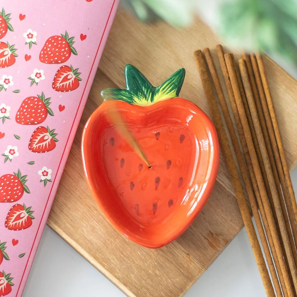 Bright red strawberry-shaped ceramic incense holder catching ash on a wooden surface with an incense stick inserted