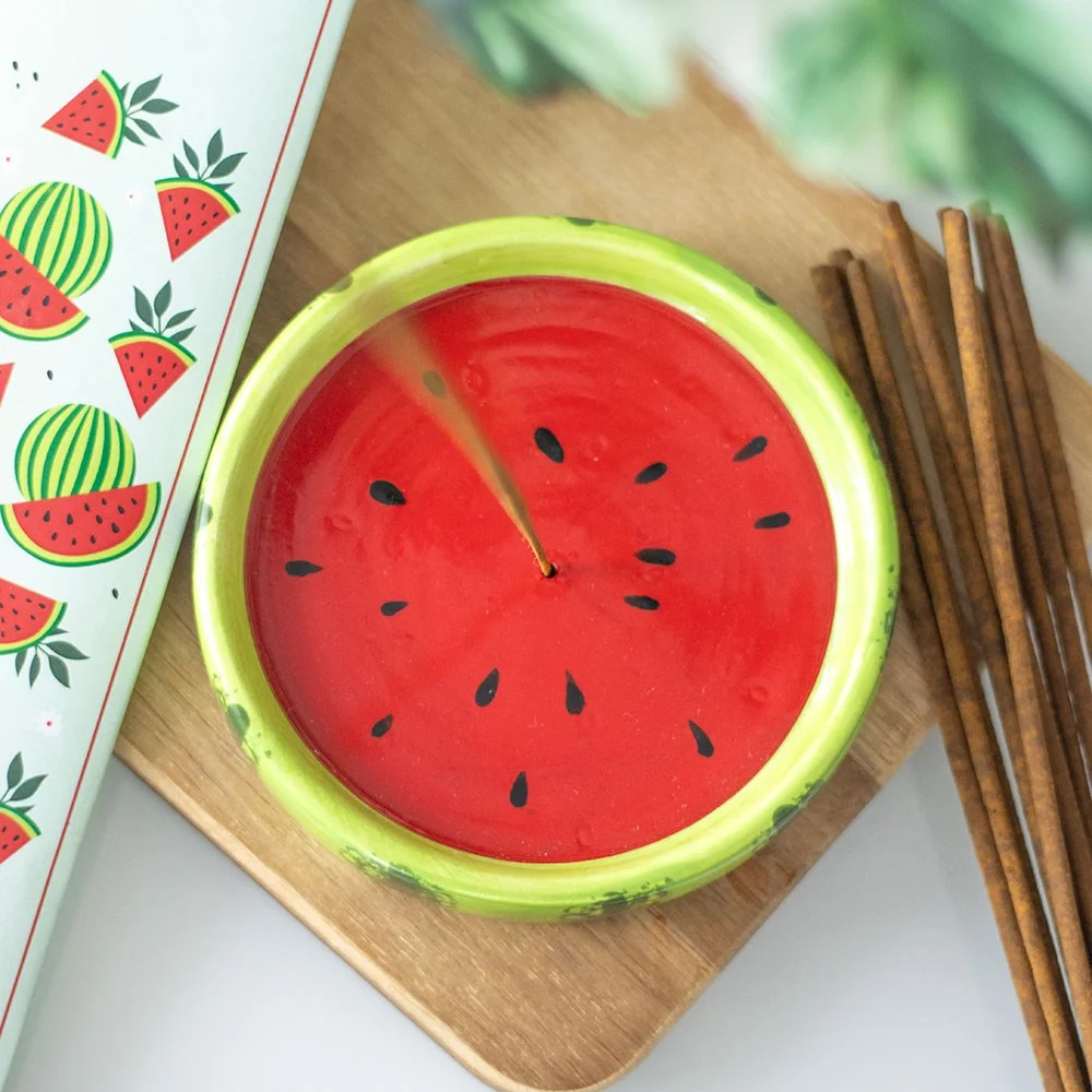 Bright ceramic incense holder shaped like a watermelon with a stick and ash catcher on a wooden surface