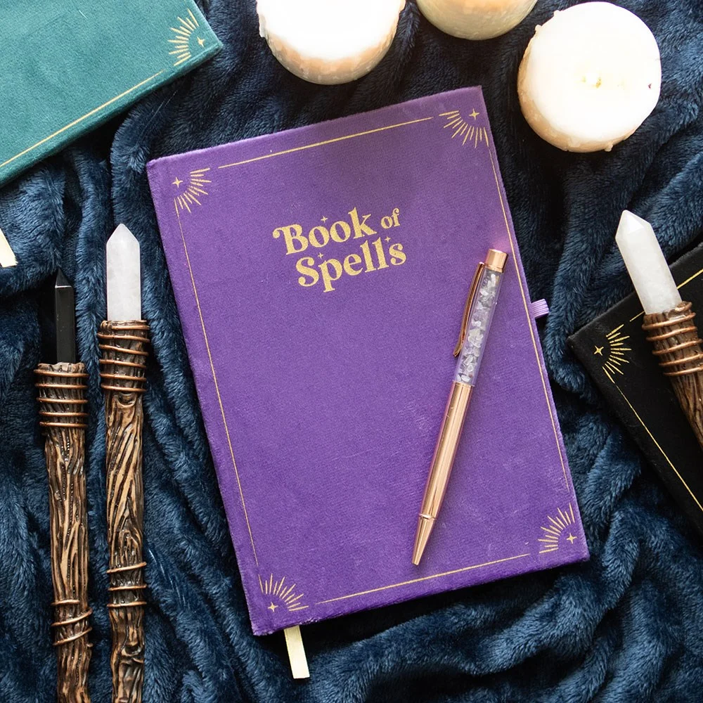 Purple Book of Spells journal with metallic gold cover details, gold ribbon marker, and amethyst crystal pen on white background