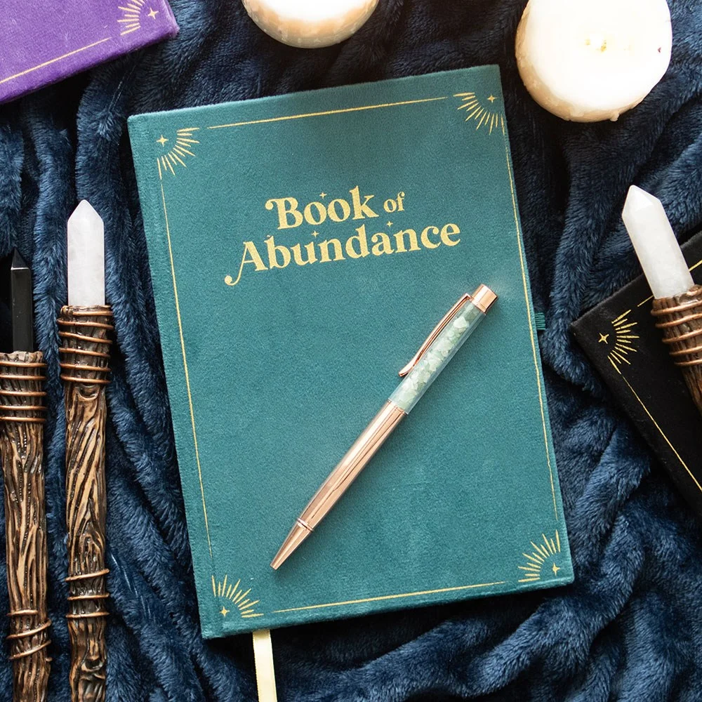 Green abundance journal with metallic gold detailing, gold ribbon page marker, and a green aventurine crystal pen beside it.