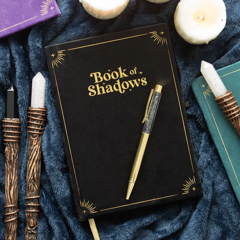 Black book of shadows journal with gold mystical detailing and ribbon marker, paired with black obsidian crystal pen on white background