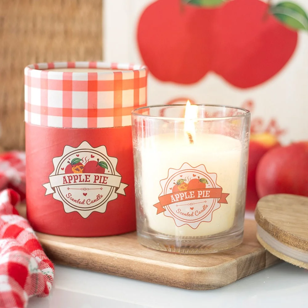 Lit apple pie scented candle in clear glass jar with warm orange label on wooden surface