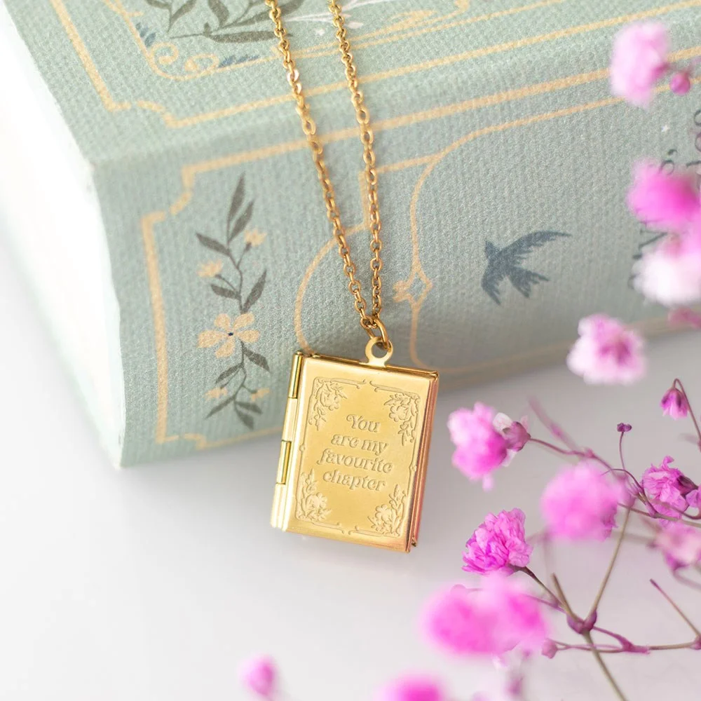 Gold-toned book shaped locket necklace in a matching gift box, open like a miniature book, showing polished stainless steel.