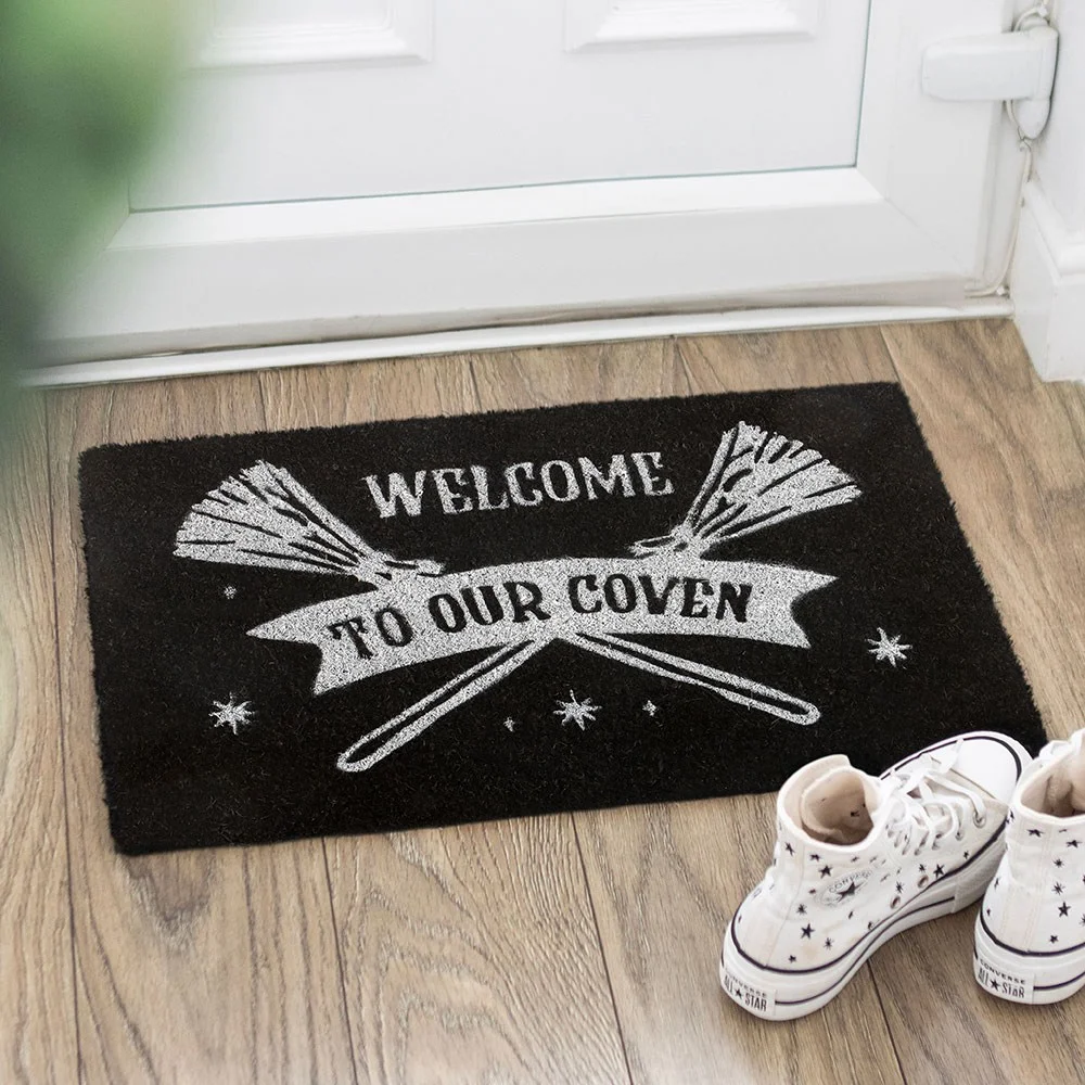 Black coven doormat made of coir, featuring silver broomsticks and a whimsical welcome message.