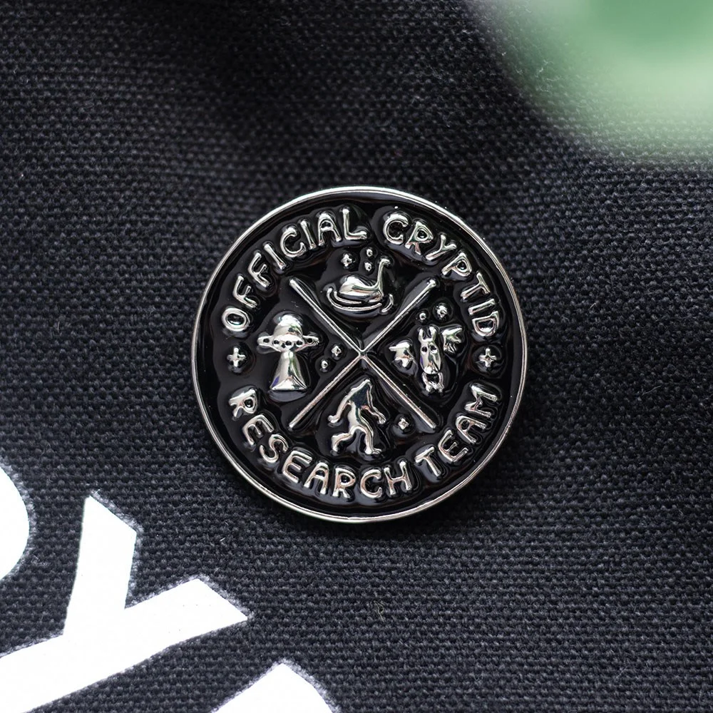 Cryptid Research Team enamel pin badge featuring a stylized Bigfoot and flying saucer design, cryptid enamel pin