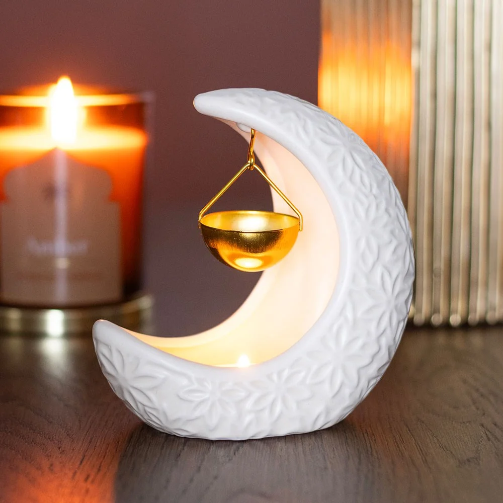 Ceramic crescent moon oil burner hanging with lit tealight candle inside, glowing warmly in dim setting