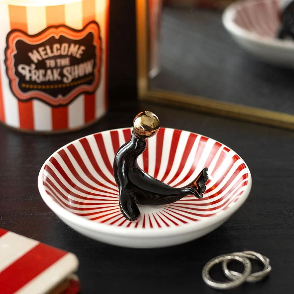 ceramic trinket dish with black circus seal balancing a golden ball on bold red and white stripes, a playful decorative piece