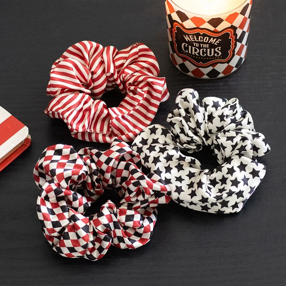 Three circus hair scrunchies in vintage star, harlequin diamond, and striped big‑top red prints, each made of 100% polyester.