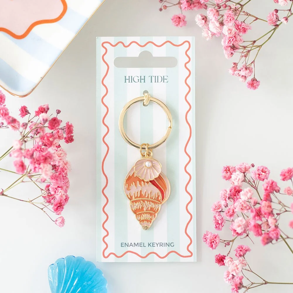 Gold-toned enamel shell keyring with soft coastal tones hanging against a neutral background