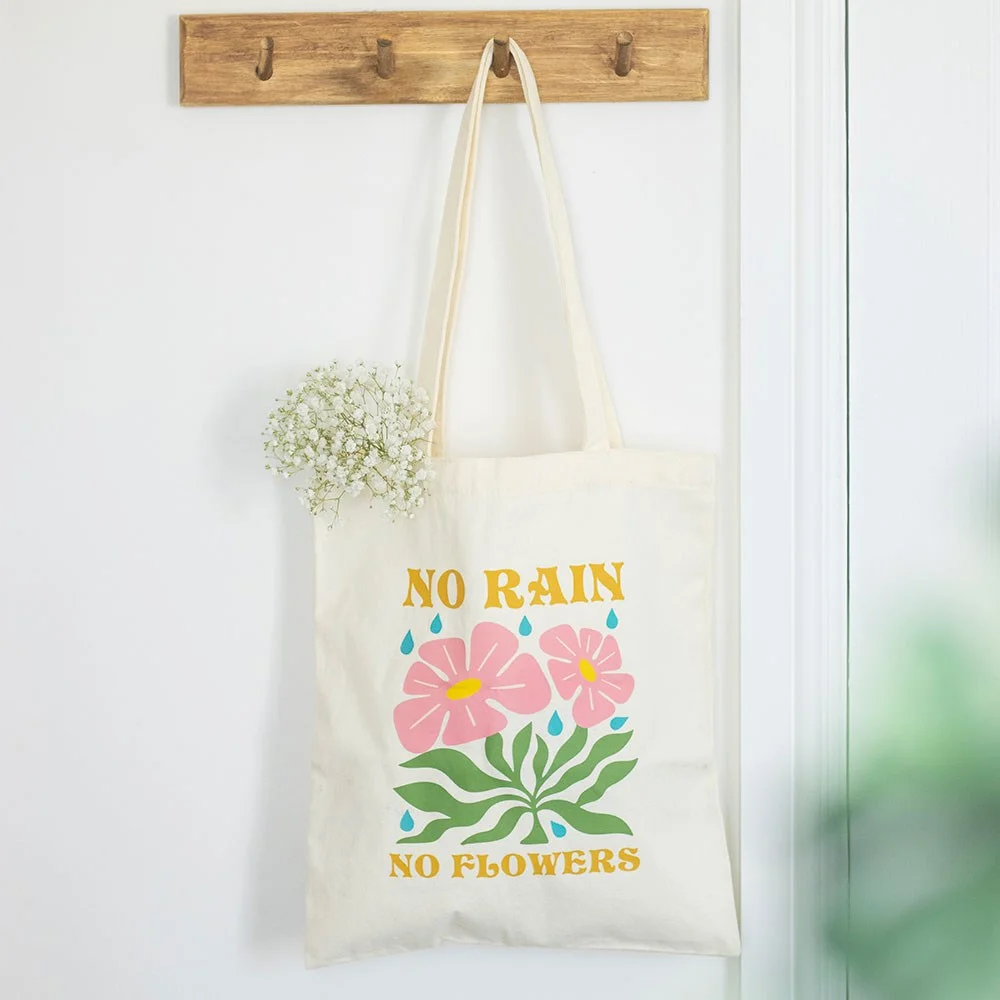 White canvas tote with printed phrase “No Rain, No Flowers”, sturdy straps, hand‑washable, no rain no flowers tote