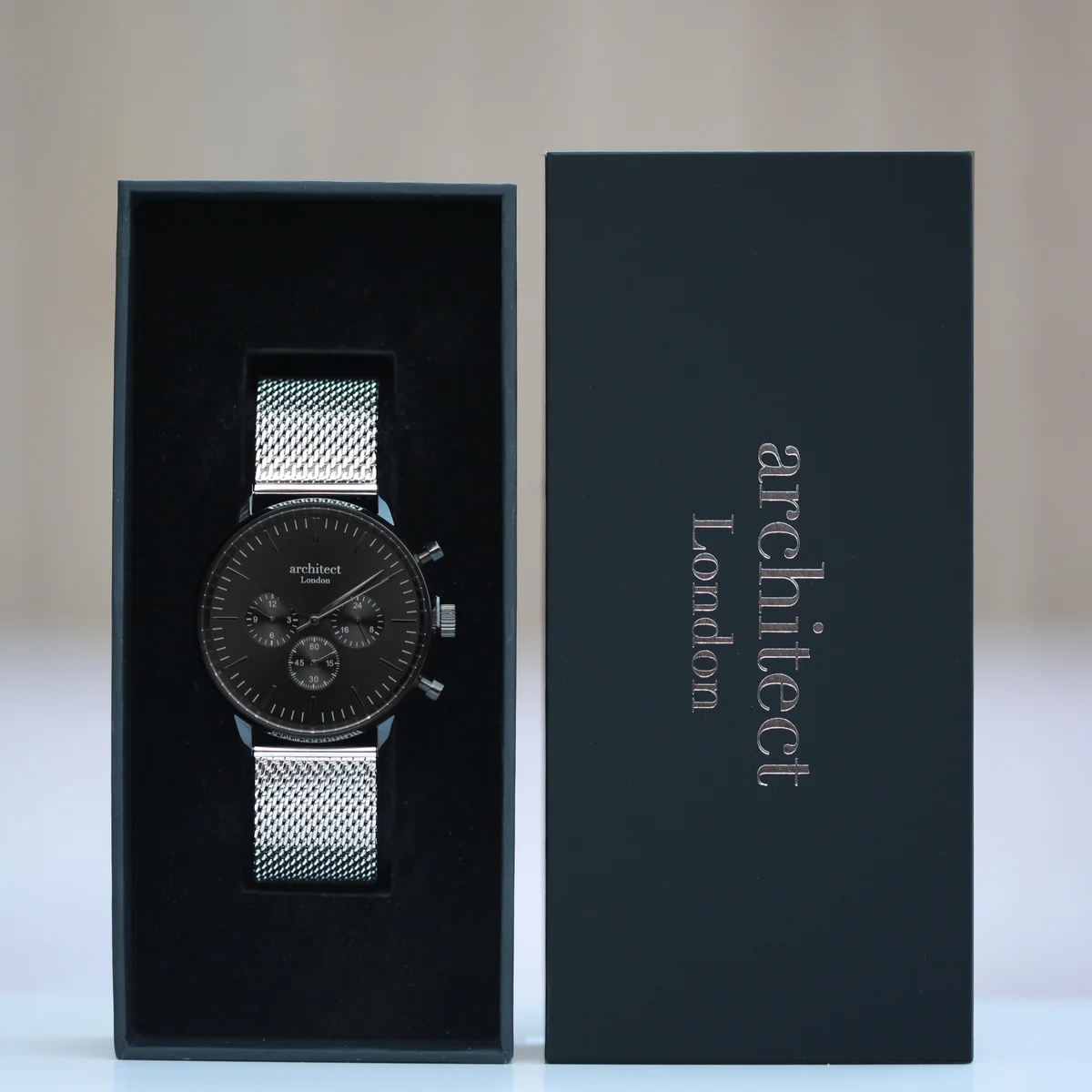 Men’s black watch with silver milanese mesh strap, chronograph dials, and modern engraved font on back case