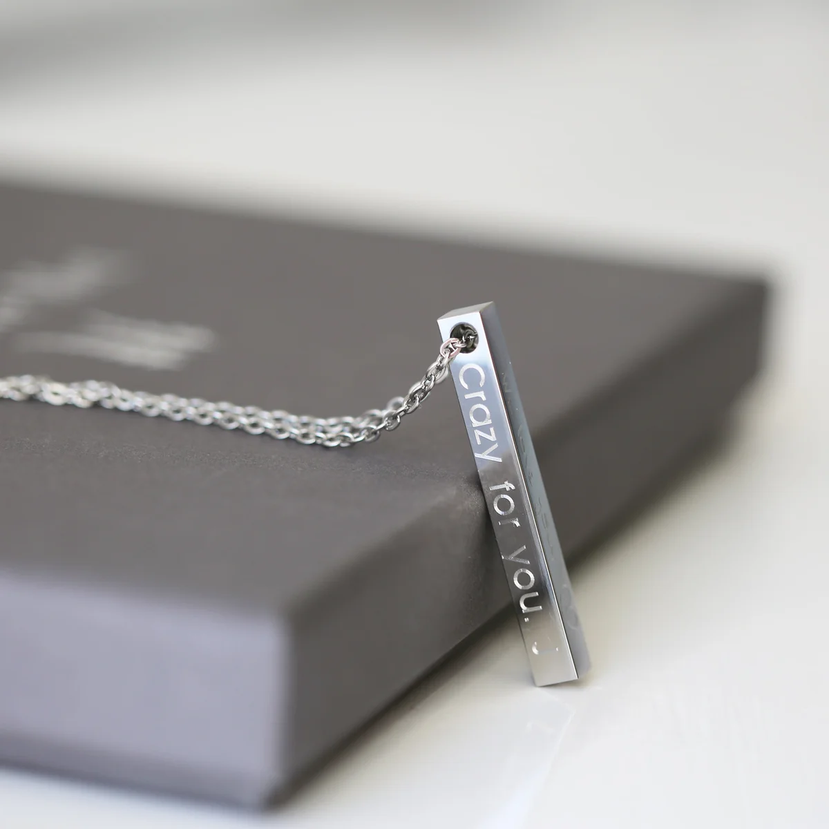 Delicate modern bar necklace in silver with engraved text, displayed on a white background with gift box nearby