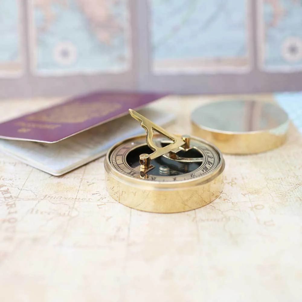 Brass sundial compass with engraved lid, showing compass face and gnomon, next to protective pouch on white background